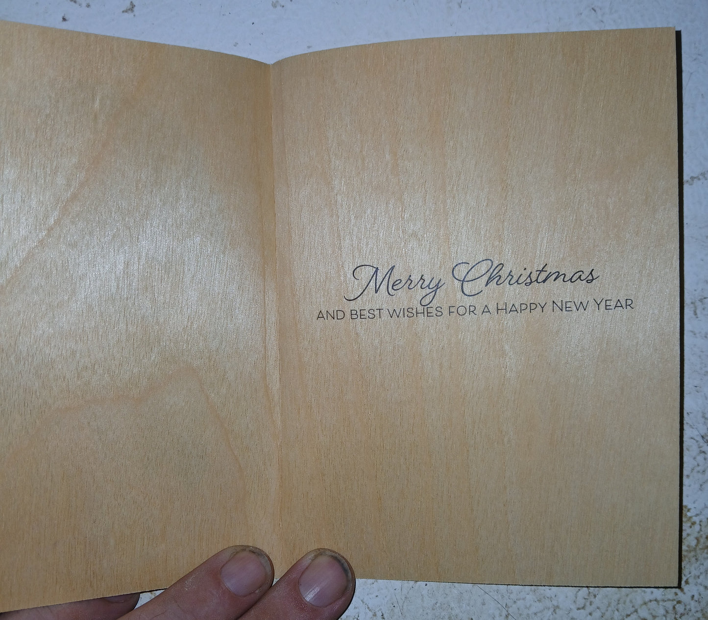 An open wooden greeting card held by a hand, showing a light natural wood interior with the printed message 'Merry Christmas AND BEST WISHES FOR A HAPPY NEW YEAR' in dark script and serif text.