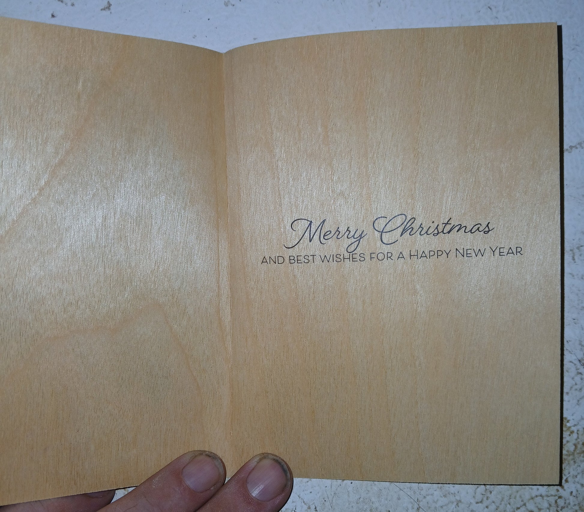 An open wooden greeting card held by a hand, showing a light natural wood interior with the printed message 'Merry Christmas AND BEST WISHES FOR A HAPPY NEW YEAR' in dark script and serif text.